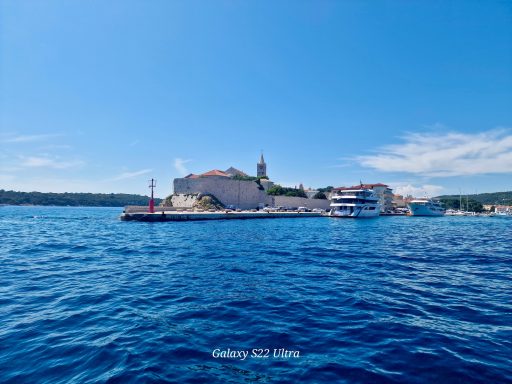 Kroatien Insel Rab Küste mit Gebäude und Kirchturm, blauer Himmel und ruhiges Wasser.