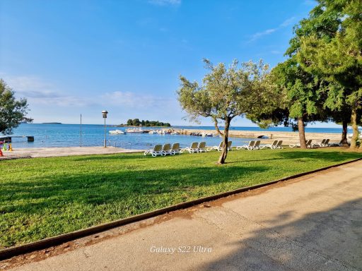 Kroatien Strandpromenade mit Liegen, Bäumen und Blick auf das Wasser und eine kleine Insel.